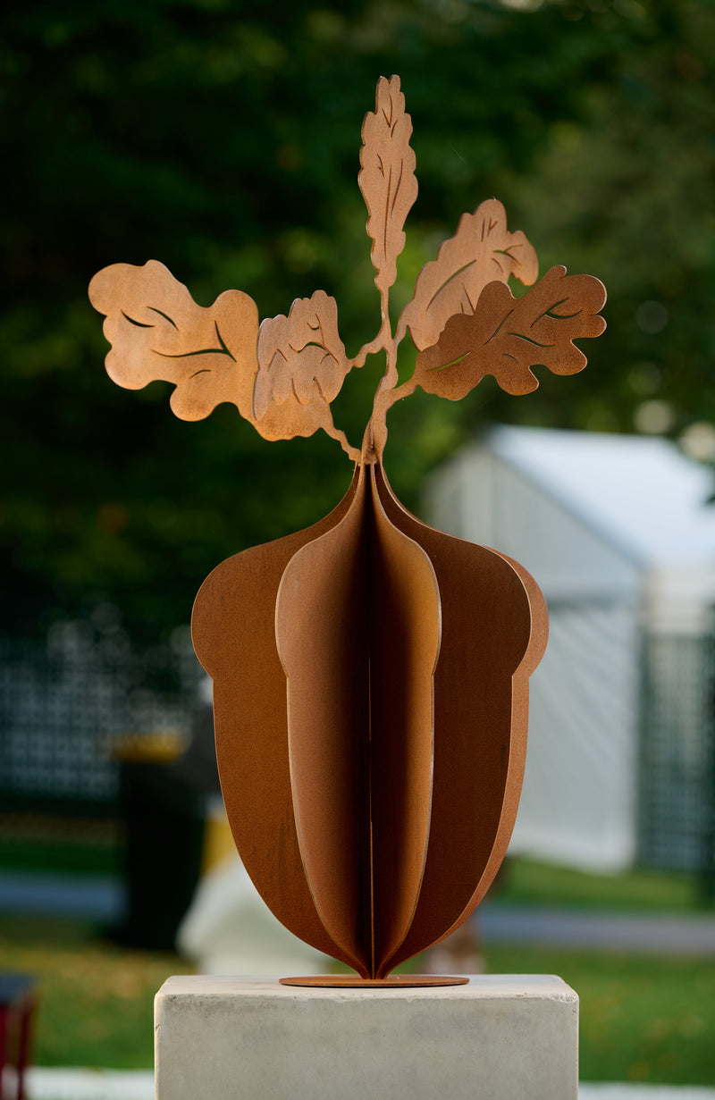 : A Corten rust 3-layer metal sculpture sitting on a concrete display stand under some tall oak trees, featuring an artistic design.  Brief Description: This image showcases a Corten rust 3-layer metal sculpture positioned on a concrete display stand. The sculpture is situated under tall oak trees, highlighting its artistic and natural design. The layers and rust finish give it a rustic and elegant appearance, blending harmoniously with the surrounding nature.