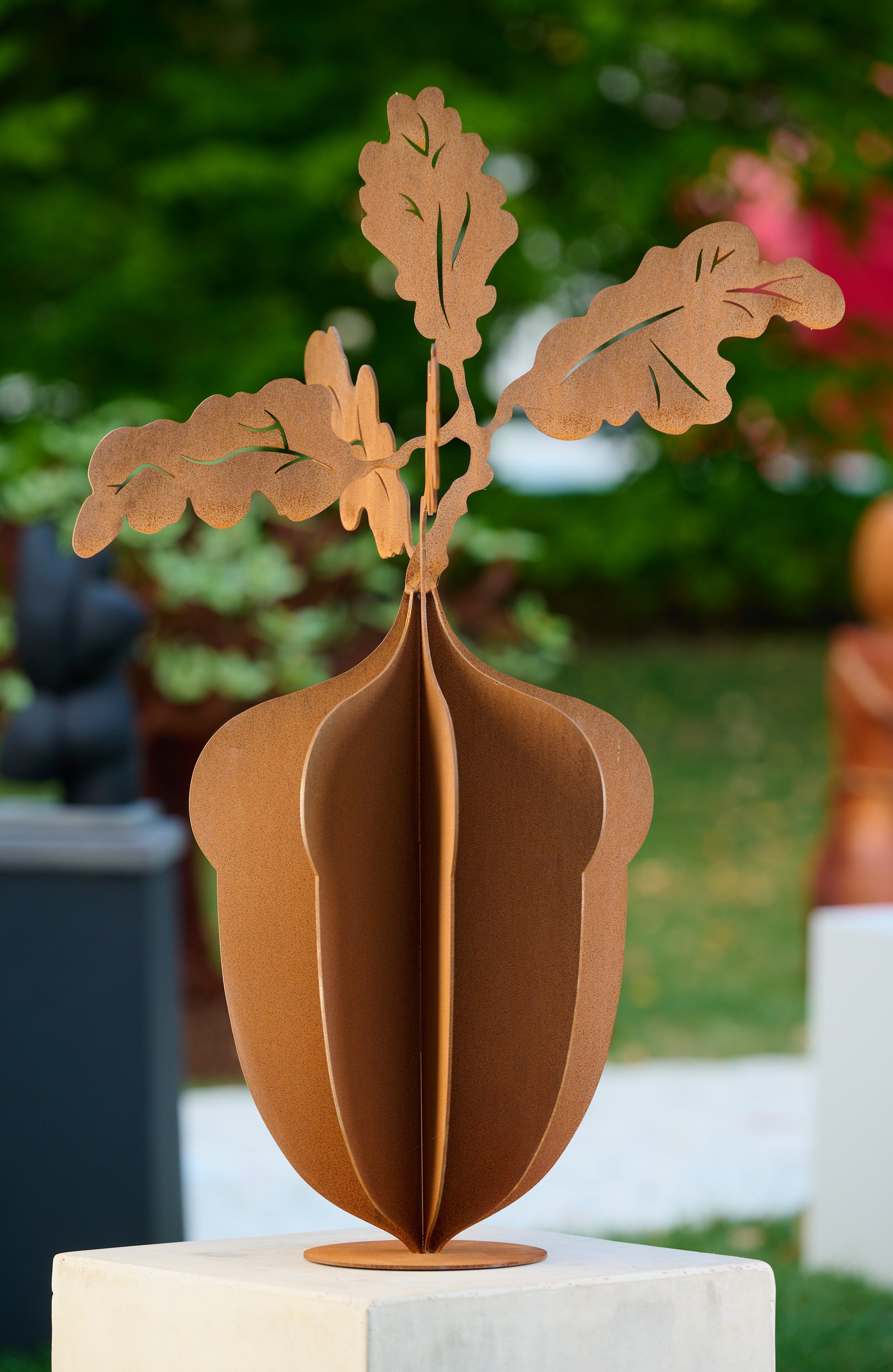 his image showcases a Corten rust 3-layer metal sculpture positioned on a concrete display stand. The sculpture, featuring an artistic design, is situated under tall oak trees. The layers and rust finish give it a rustic and elegant appearance, blending harmoniously with the surrounding nature.