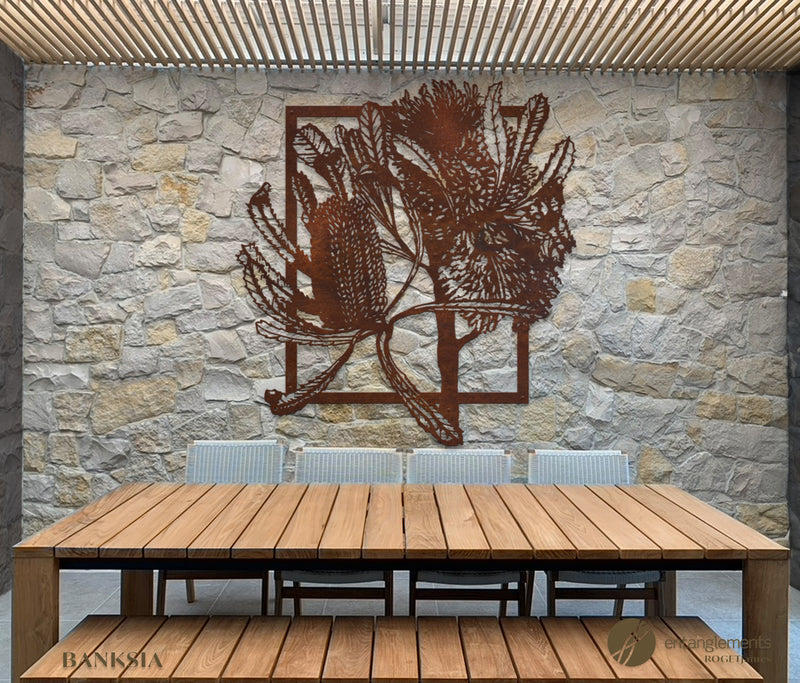  shows a rustic metal wall art piece featuring a silhouette of banksia. The art is set against a crazy-paved light sand stone wall, with the scene situated in a light timber alfresco dining area. The contrast highlights the silhouette of the banksia against the textured background.